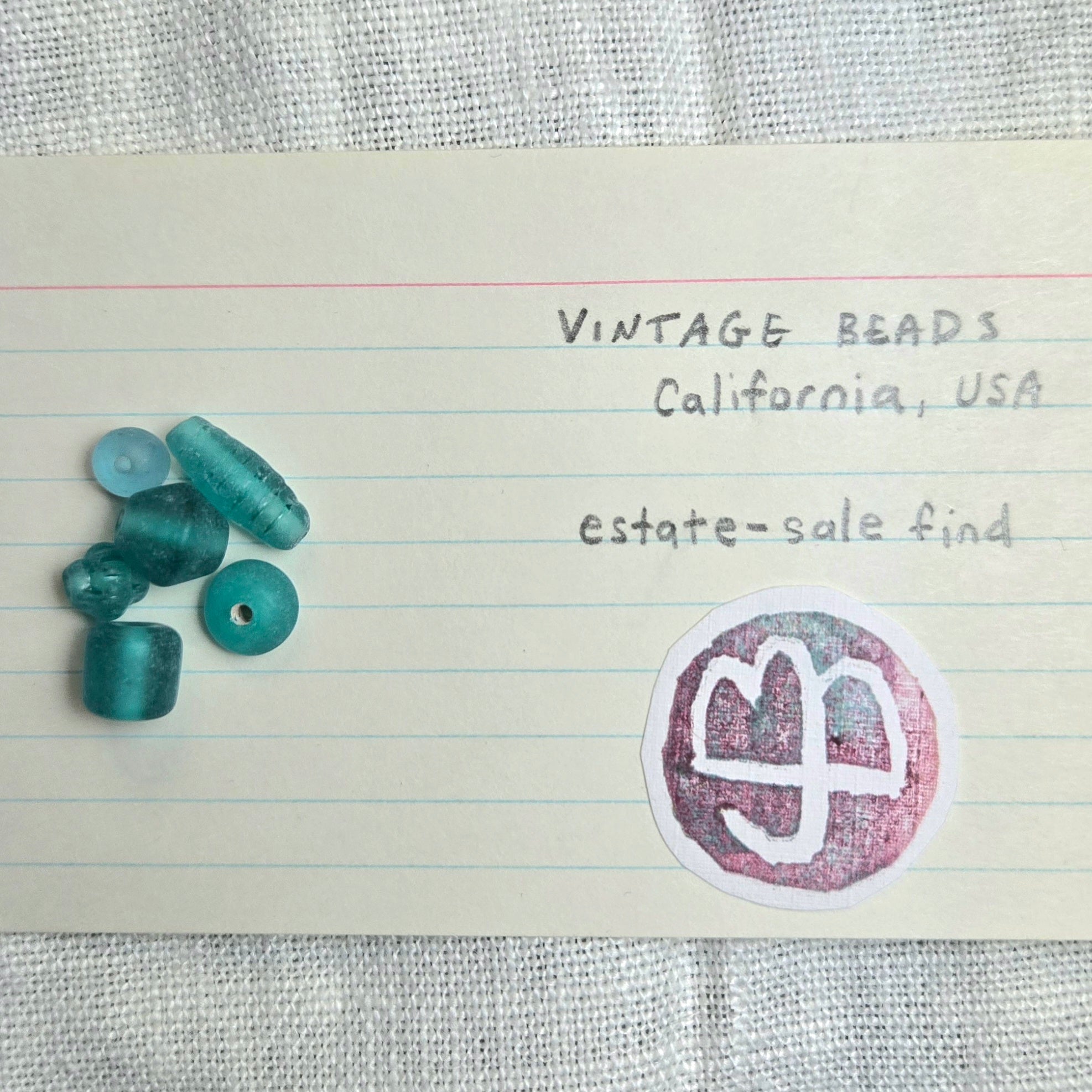 A set of vintage teal glass beads arranged on a piece of lined notebook paper next to handwritten text reading 'VINTAGE BEADS California, USA estate-sale find' and a circular stamp.
