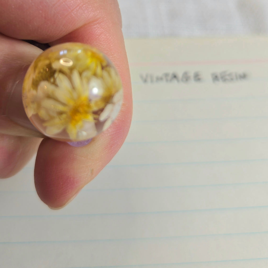 A close-up of a hand holding a round clear resin bead containing dried yellow and white flowers, with lined paper in the background that has the handwritten words 'VINTAGE RESIN'.