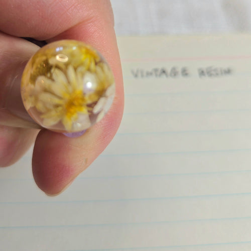 A close-up of a hand holding a round clear resin bead containing dried yellow and white flowers, with lined paper in the background that has the handwritten words 'VINTAGE RESIN'.