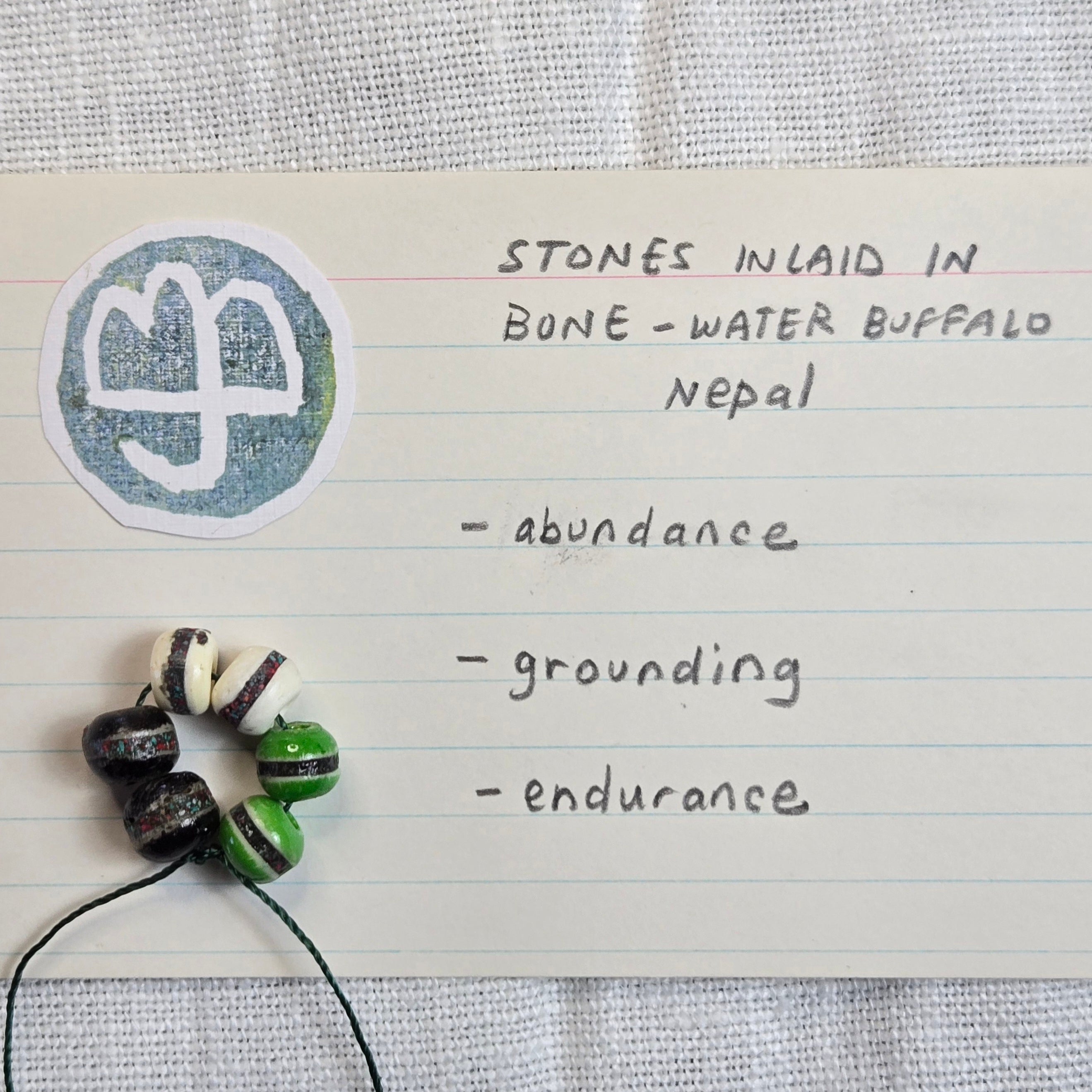 A handmade beaded necklace with black, white, and green patterned beads on a black cord, placed on a piece of lined paper next to a circular sticker with a white symbol. The paper has handwritten text reading 'STONES INLAID IN BONE - WATER BUFFALO NEPAL - abundance - grounding - endurance'.