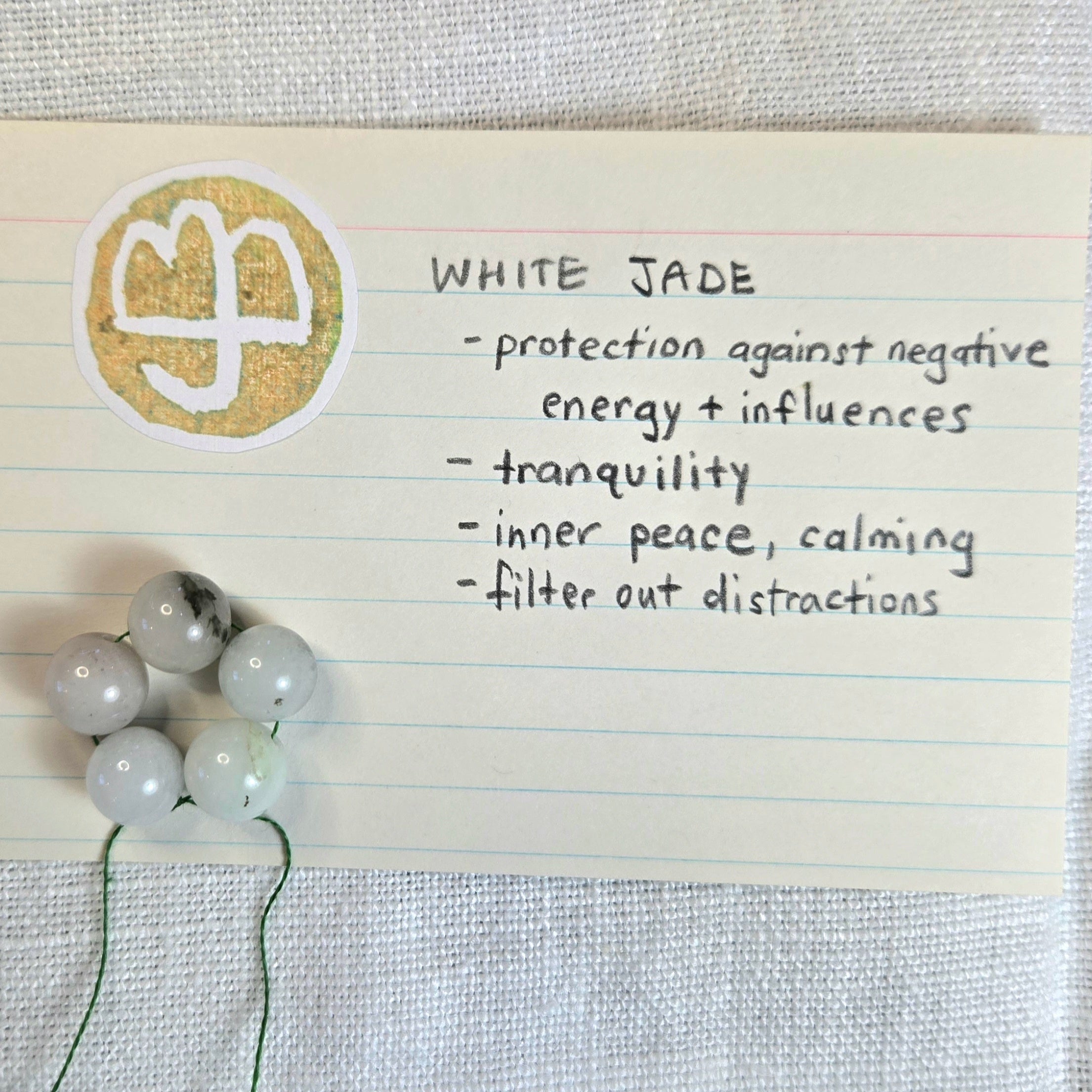 A handwritten note on lined paper identifies the item as white jade, listing its purported benefits. Several white jade beads with dark inclusions are threaded on a green string, placed next to the note on a textured white fabric background.