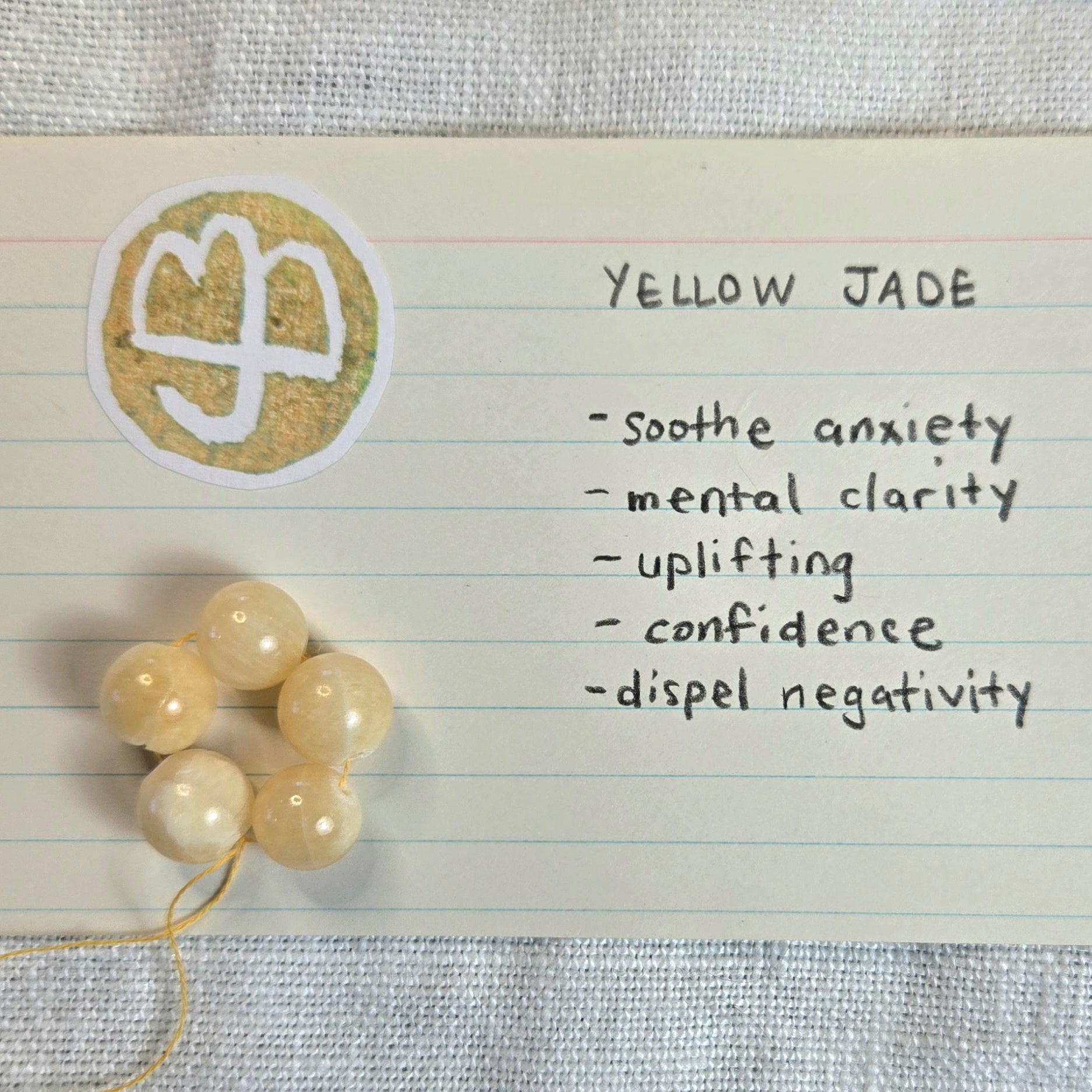 A small card labeled 'YELLOW JADE' with handwritten benefits and a circular symbol, next to a cluster of pale yellow jade beads on a textured fabric background.